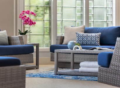 A bright, cozy outdoor seating area with blue and gray cushions, a wicker chair, a coffee table, potted plant, and a sunny view through large windows.