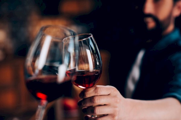 A man is holding a glass of red wine in a cozy, dimly-lit setting, with another glass of wine visible in the foreground.