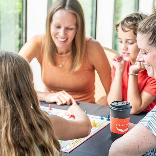 A group of people, including children and adults, are playing a board game happily indoors near large windows.