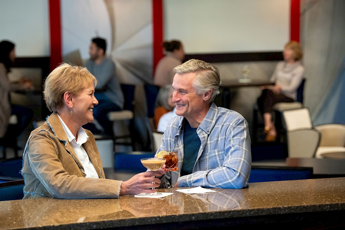 A couple is sitting at a bar, smiling and toasting with drinks, in a relaxed, casual setting with others in the background.