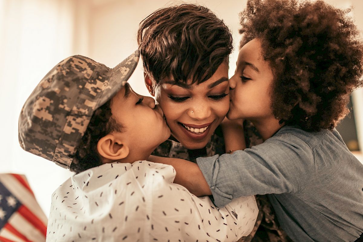 A person in a military uniform is being kissed on both cheeks by two children, one wearing a camouflage hat. They are all smiling happily.