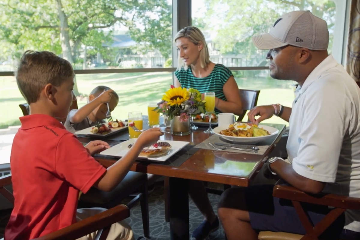A family of four is sitting at a table, enjoying a meal together in a bright, spacious setting with greenery outside the window.