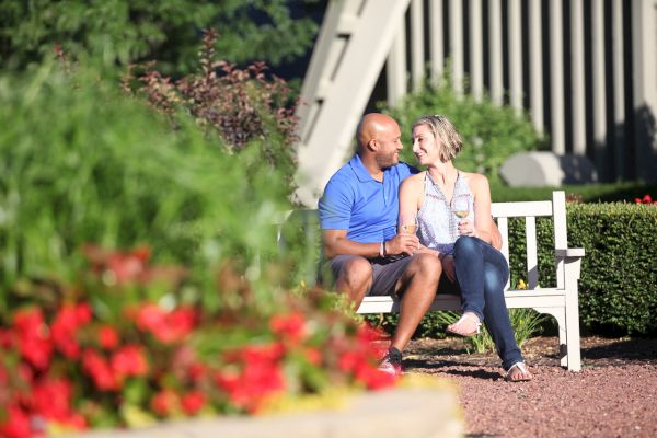A couple sits on a white bench in a garden, with colorful flowers in the foreground and a building structure in the background.