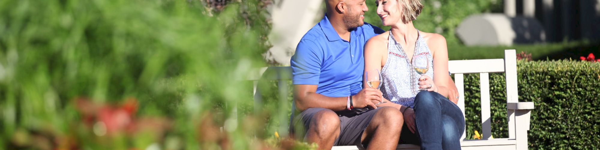 A couple sits closely on a white bench, holding drinks, surrounded by greenery and red flowers in a garden setting.