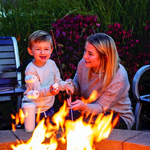 A woman and a boy enjoy roasting marshmallows by a backyard fire pit at dusk, sharing a joyful moment together.
