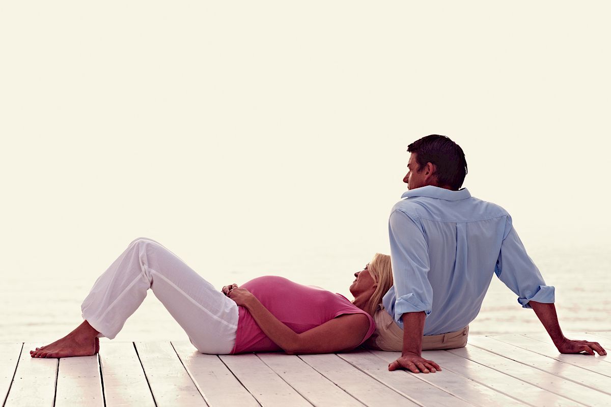 A pregnant woman in a pink top and white pants leans against a man in a blue shirt, both sitting on a wooden deck.