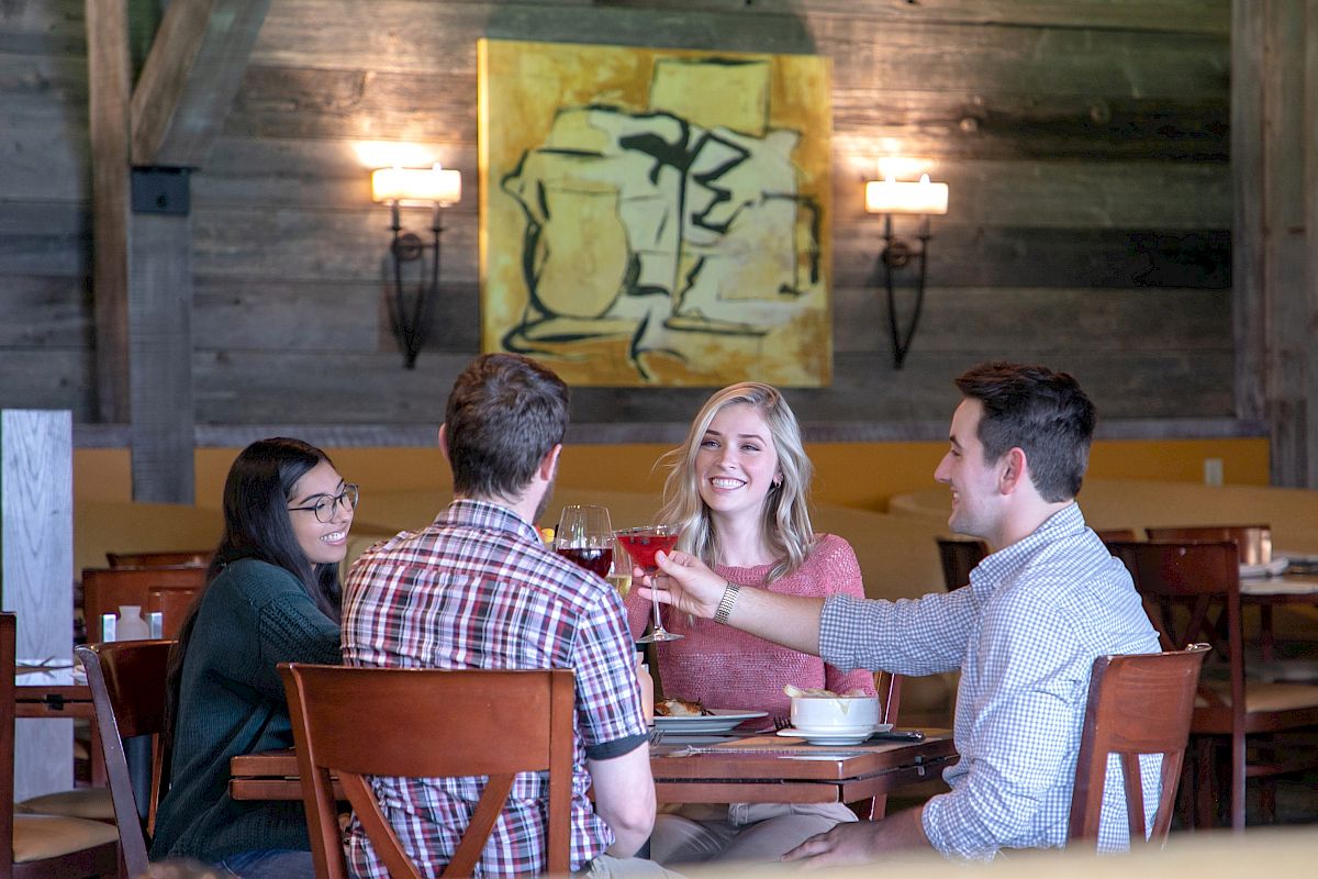 Four people are sitting at a restaurant table, smiling and toasting with drinks. There is abstract artwork on the wall behind them.