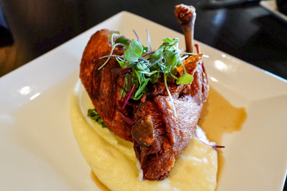 A roasted chicken leg served on creamy mashed potatoes, garnished with microgreens on a square white plate.