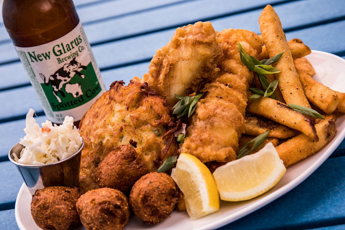The image shows a plate with fried fish, french fries, cole slaw, hush puppies, lemon wedges, and a bottle of New Glarus beer on a blue table.