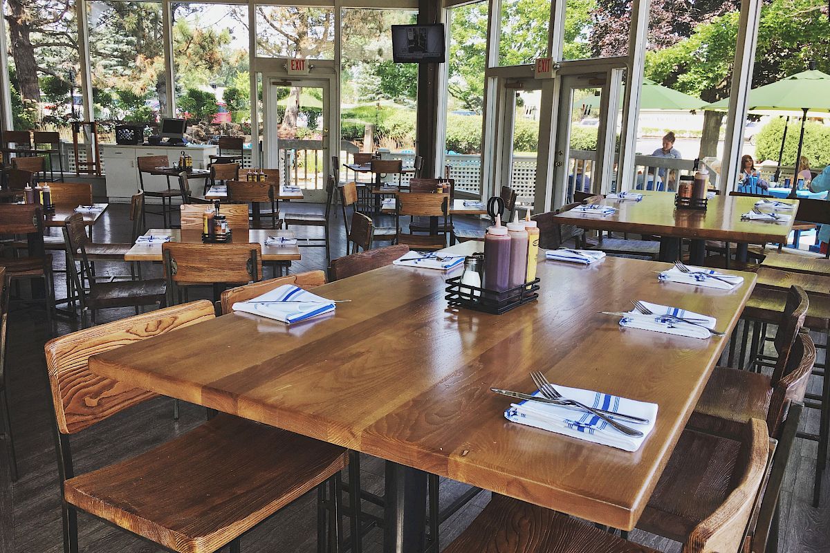 A restaurant with wooden tables and chairs, set with napkins and cutlery, and featuring large windows with a view of an outdoor seating area.