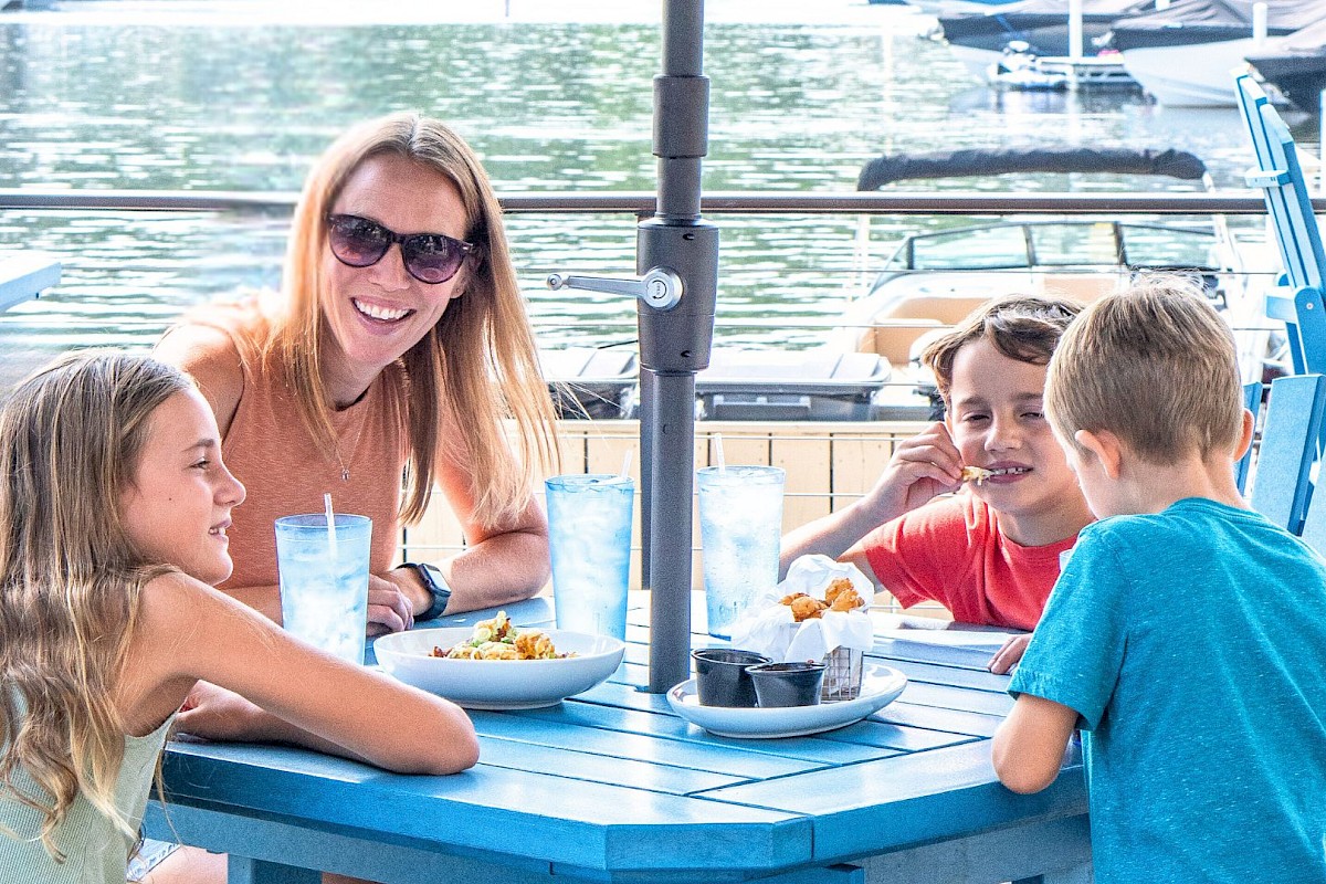 A group of people enjoying a meal outdoors at a table by the water, with boats in the background and sunny weather.