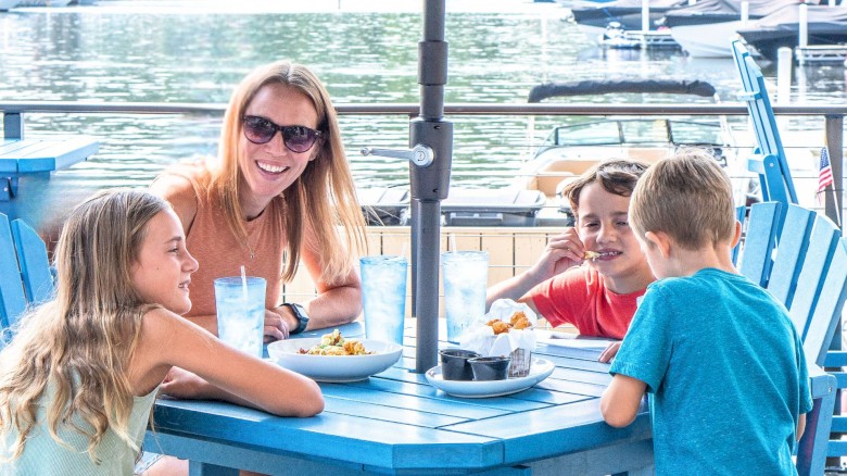 A group of people enjoying a meal outdoors at a table by the water, with boats in the background and sunny weather.