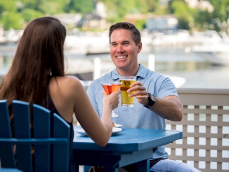 Two people clink glasses while seated at an outdoor table with a scenic waterfront view in the background.