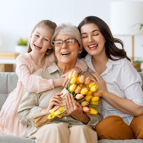 Three generations of women are smiling on a couch, with the grandmother holding a bouquet of yellow tulips.