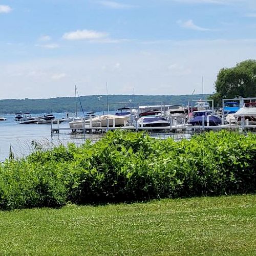 A lake scene with boats docked at a marina, lush green grass, and a bench in the foreground, under a partly cloudy sky ending the sentence.
