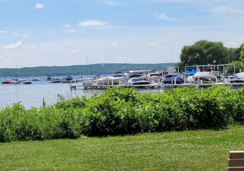 A scene of a lake with boats docked, green foliage in the foreground, clear skies, and a park bench on the right.