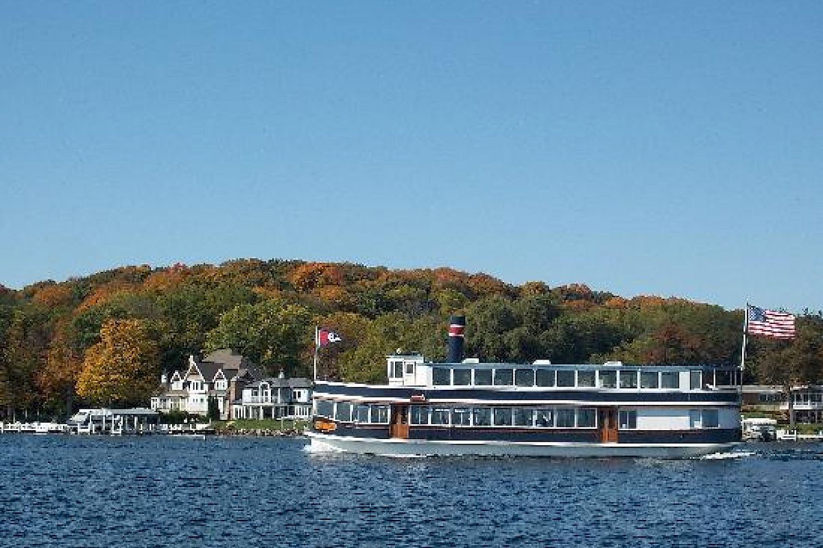 A large boat is cruising on a lake with a forested shoreline and some houses in the background, under a clear blue sky.