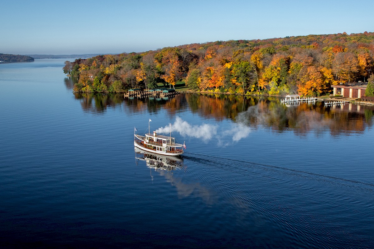 A boat sails on a calm river near a shoreline with colorful autumn trees and some houses, under a clear blue sky.