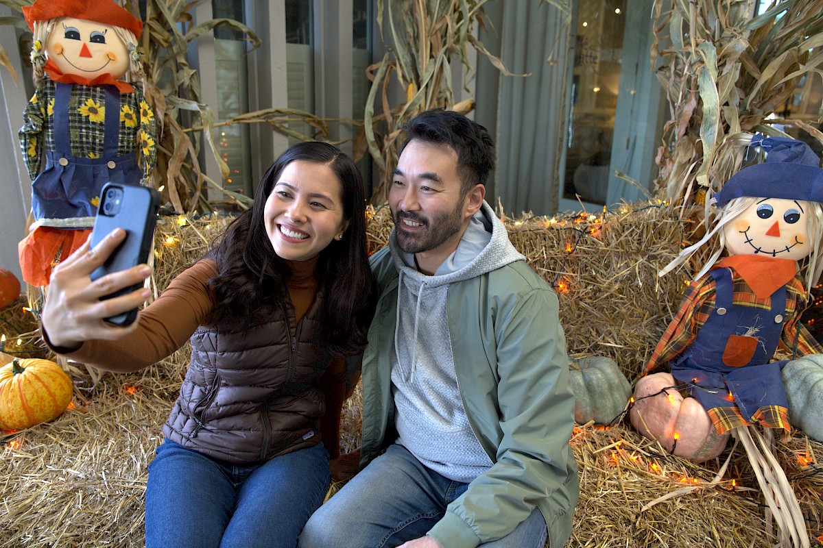 A couple taking a selfie in a festive fall setting with scarecrows, pumpkins, and straw decorations around them.