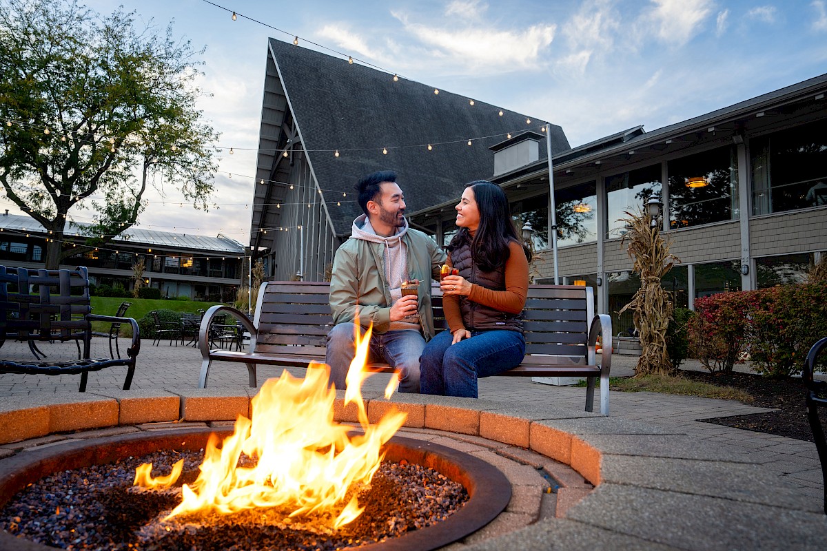 A couple sits by a fire pit, holding drinks, in an outdoor setting with string lights and a building in the background.