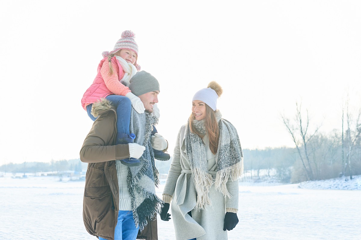 A family of three, warmly dressed, enjoying a snowy day. The child is on the father's shoulders while the mother walks beside them, holding hands.