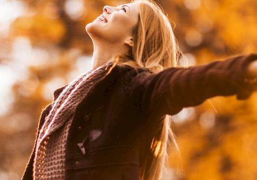 A person is outdoors with arms outstretched, enjoying the autumn scenery with a happy expression. They are wearing a scarf and a jacket.