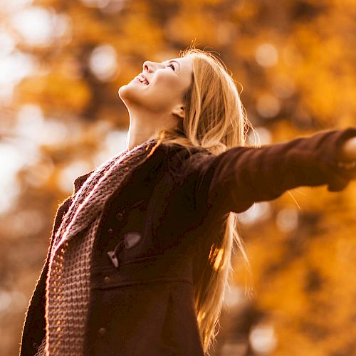 A person is outdoors with arms outstretched, enjoying the autumn scenery with a happy expression. They are wearing a scarf and a jacket.