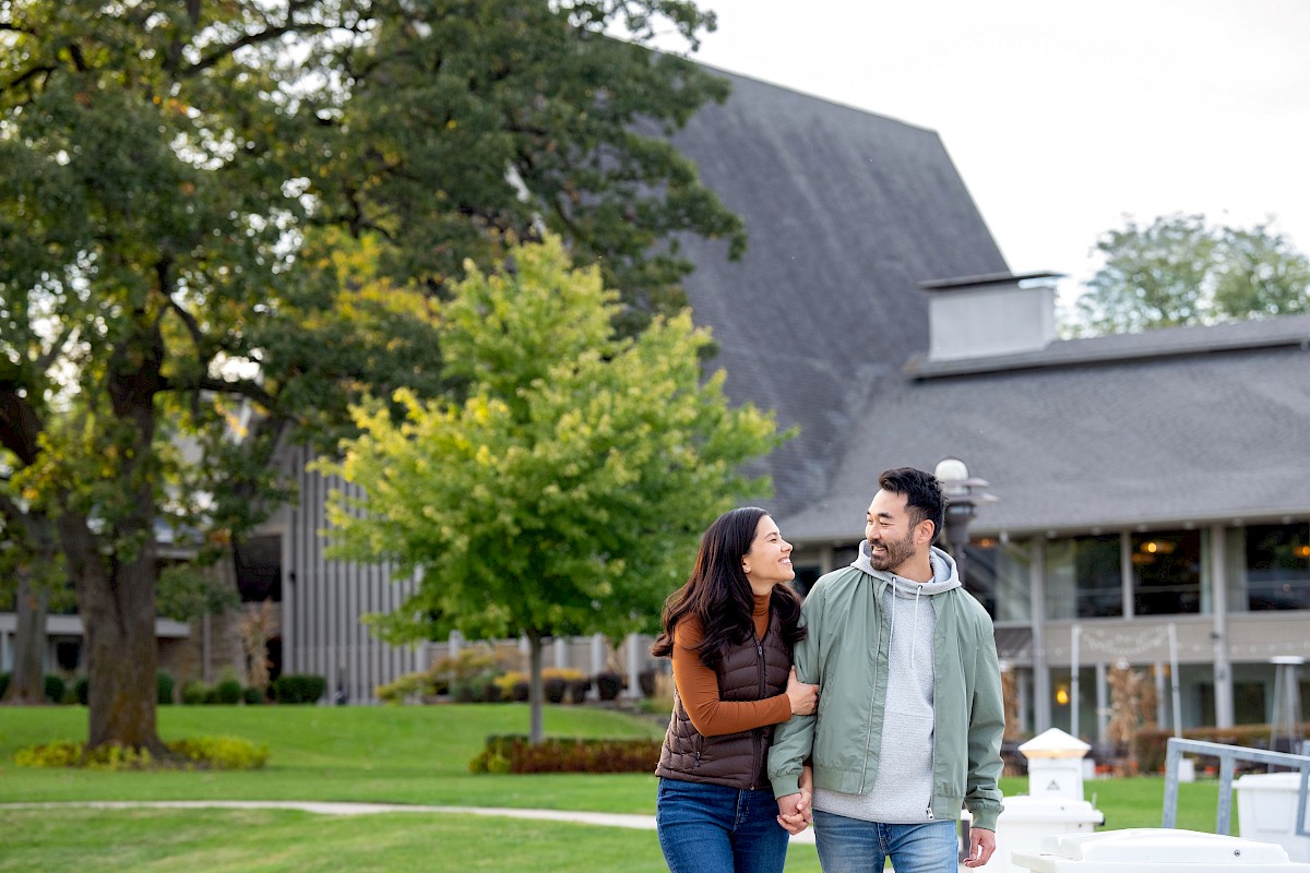 A couple happily walking hand in hand outdoors near a house with trees and green grass in the background.