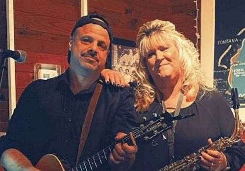 Two musicians are posing indoors; one is holding a guitar, and the other is holding a saxophone.