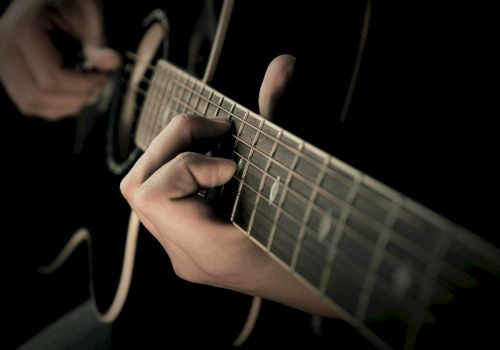 A close-up image of a person playing an acoustic guitar, showing their hands pressing the strings along the fretboard.