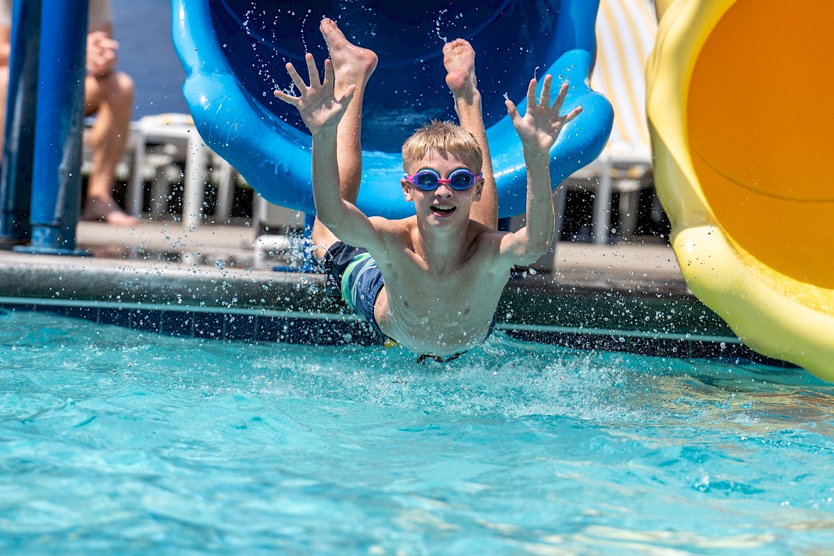 A boy with goggles is sliding out from a water slide into a pool, smiling and enjoying a sunny day at the water park.