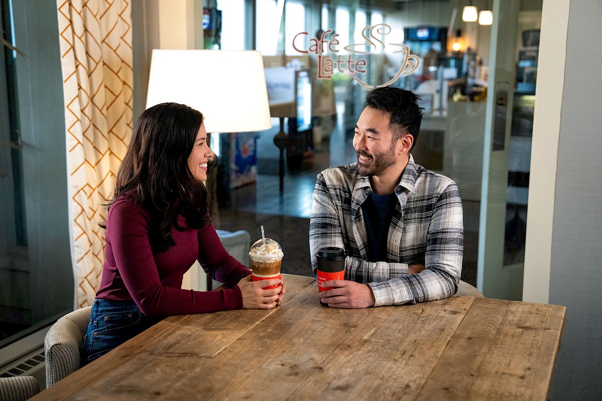 A man and woman sit at a table in a cafe, smiling and holding drinks, with the cafe's logo visible in the background.