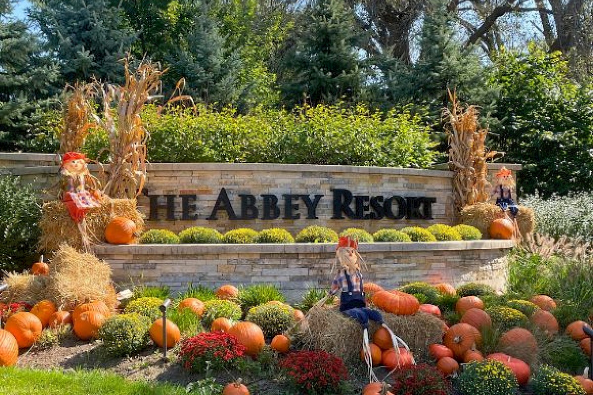 The image shows an autumn-themed entrance with pumpkins, scarecrows, and fall decorations in front of a sign that reads 