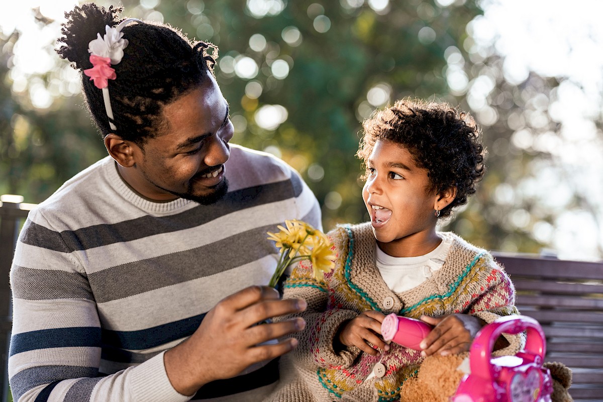 A man and a young girl share a joyful moment outdoors, with the man giving her flowers as they smile at each other.