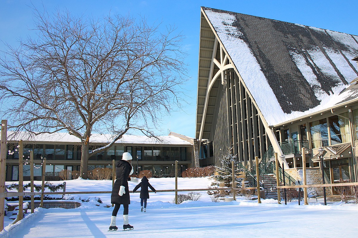 Two people skate on a snowy outdoor rink beside a modern, triangular-roof building in winter sunshine.