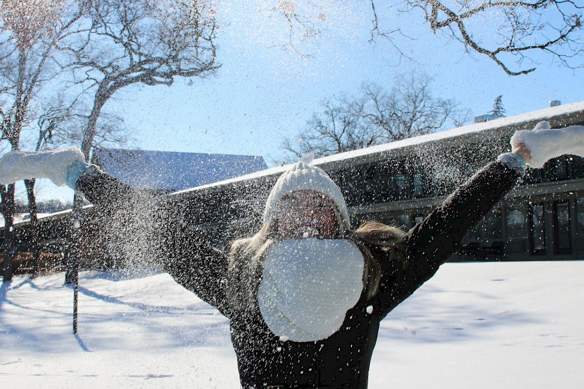 A person in black winter clothing with arms outstretched enjoying snow, wearing a white hat and face mask, in a snowy outdoor scene.