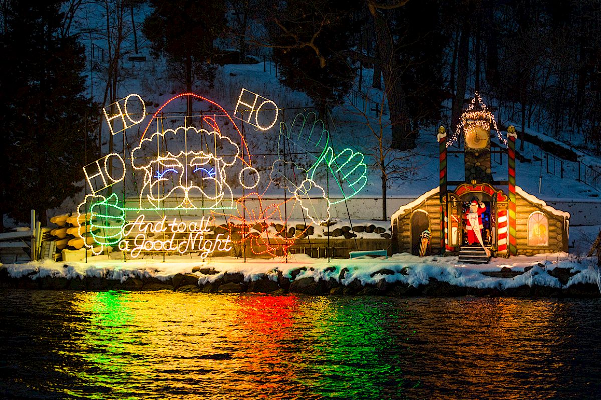 A festive holiday display featuring Santa Claus lights and a decorated house, reflected on a water surface, in a snowy landscape, ending the sentence.