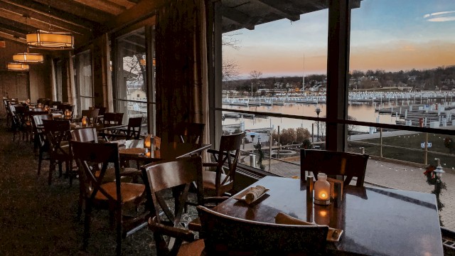 A chic, dimly lit restaurant interior with wooden chairs and tables, large windows overlooking a marina at sunset. End.