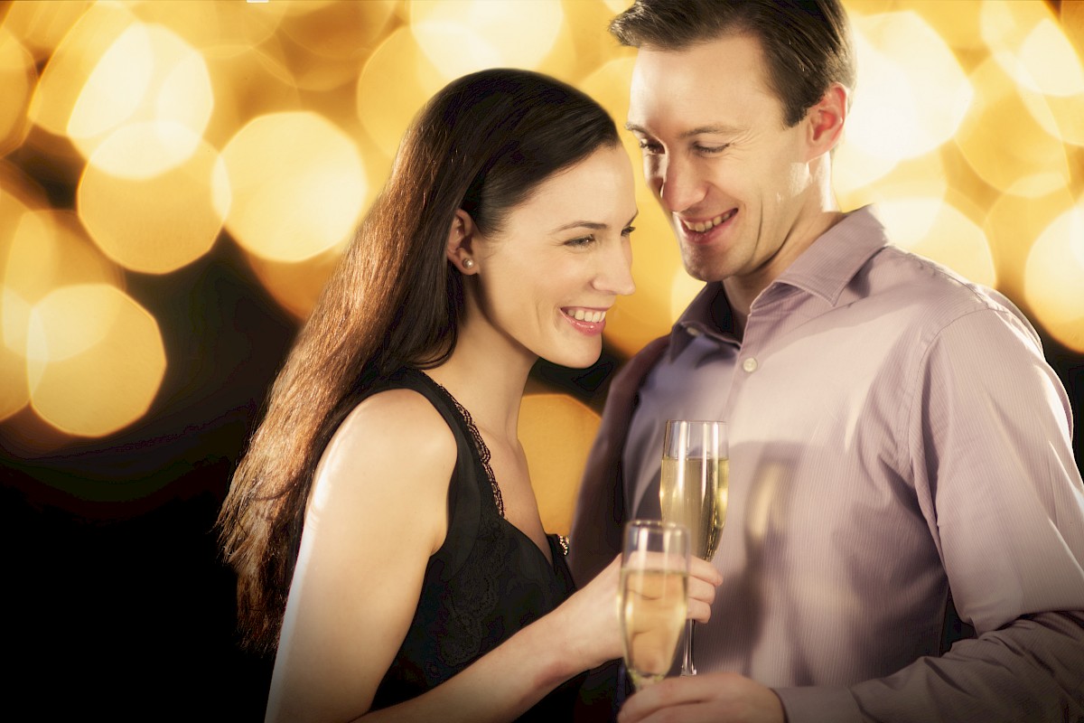 A couple is smiling and holding champagne glasses, with a warm, blurry background of golden lights.