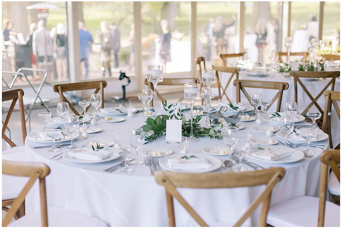 A round table set for a formal event with white tablecloth, greenery centerpiece, glassware, and wooden chairs.