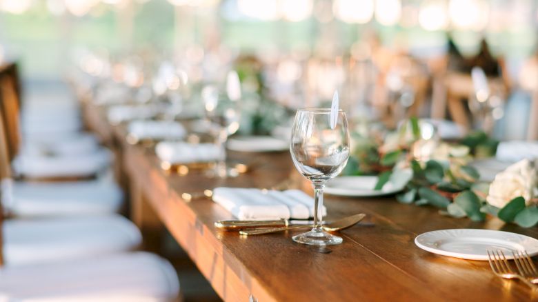A long wooden table is set for a formal event, with white plates, silverware, wine glasses, and floral decorations.