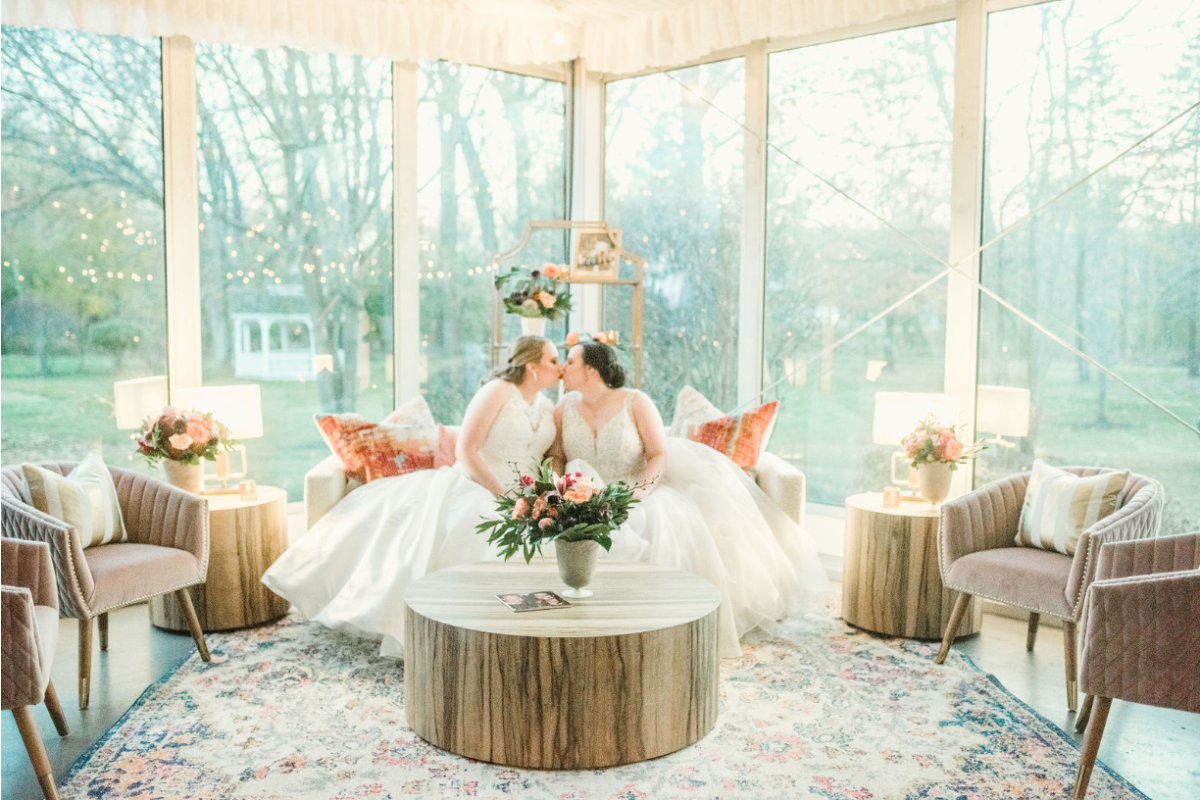 Two brides in white dresses share a kiss in a beautifully decorated room with large windows, cozy seating, and floral arrangements.