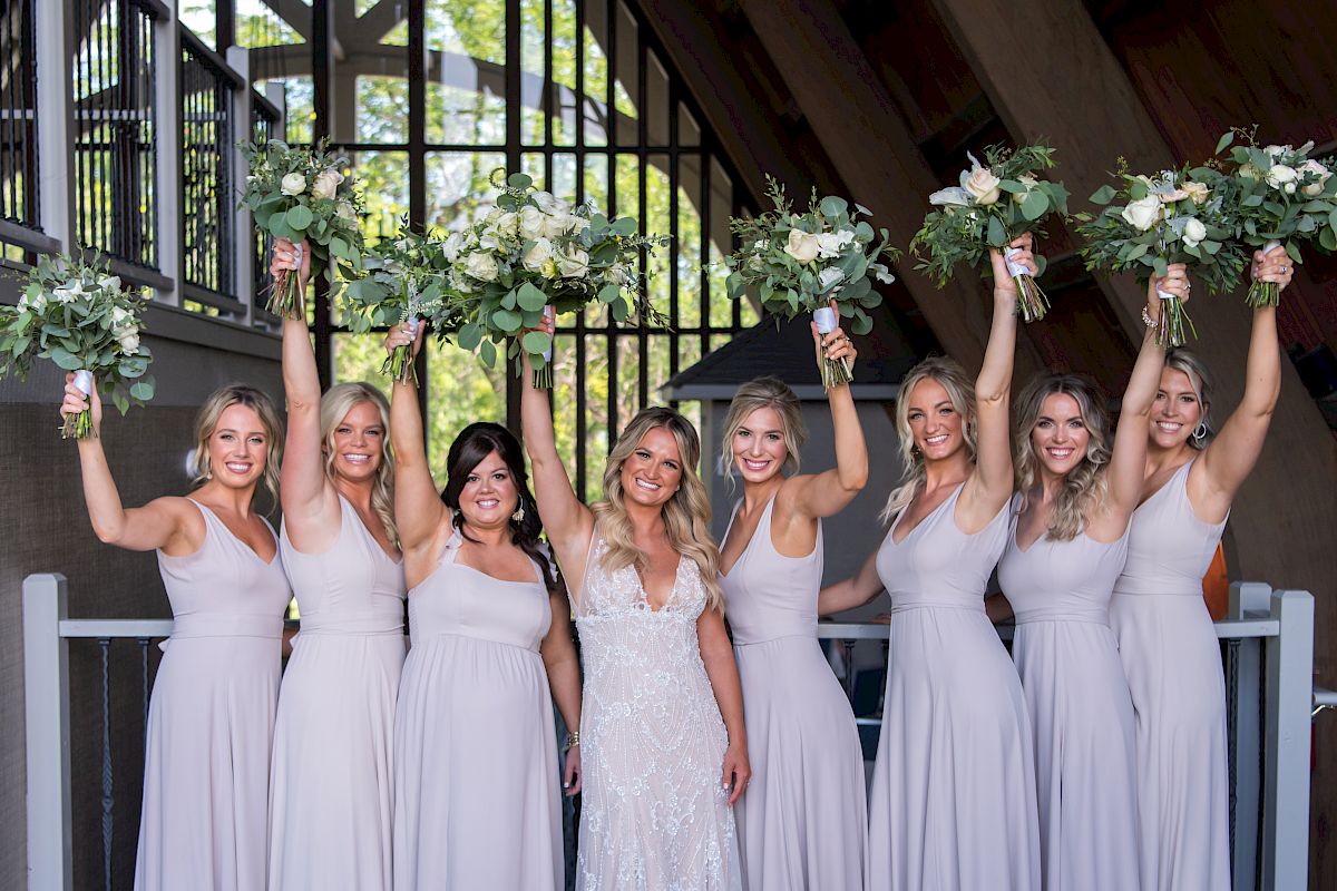 A bride stands center, surrounded by six bridesmaids in lavender dresses, all holding bouquets aloft, in an airy venue with natural light.