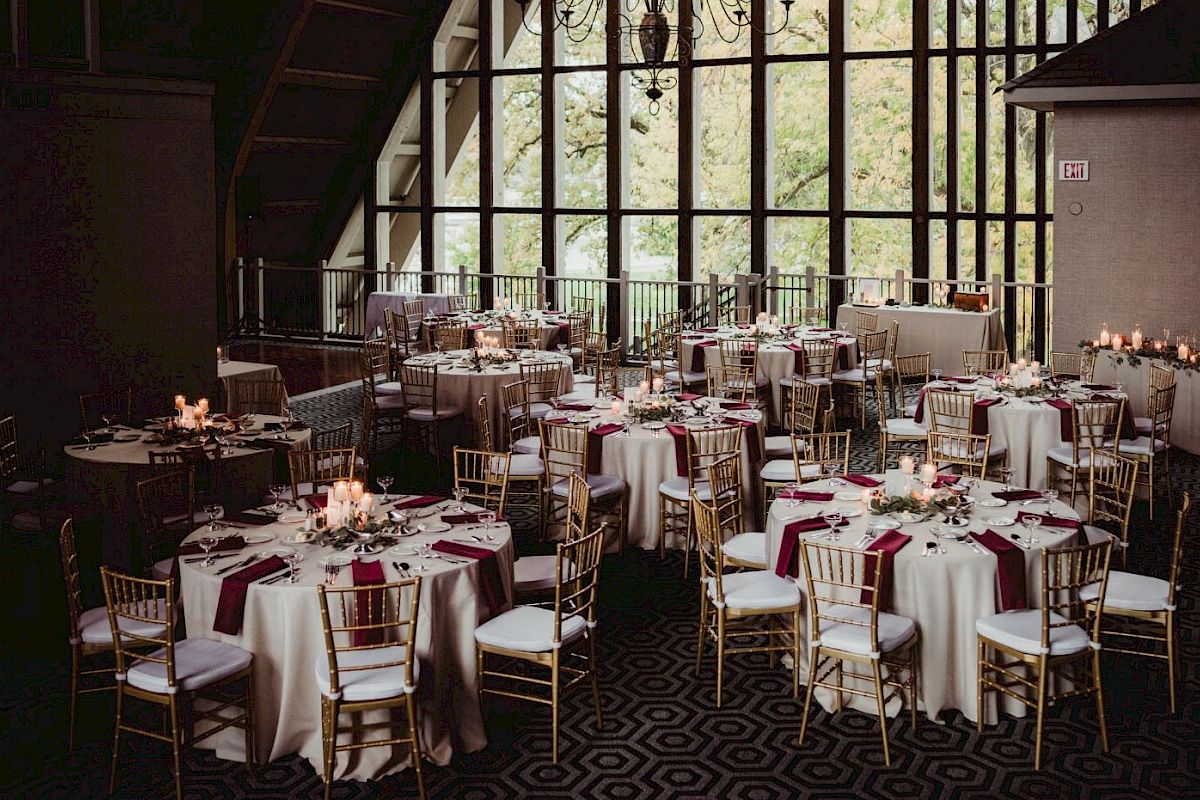 A decorated event space with round tables, white tablecloths, red napkins, and gold chairs, ready for a gathering or celebration, with candles on tables.