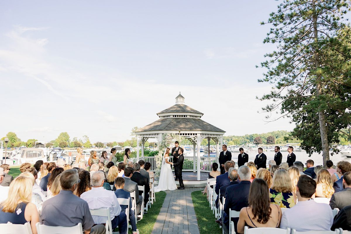 A wedding ceremony in progress outdoors by a gazebo with a crowd seated on both sides, the couple exchanging vows in the center.