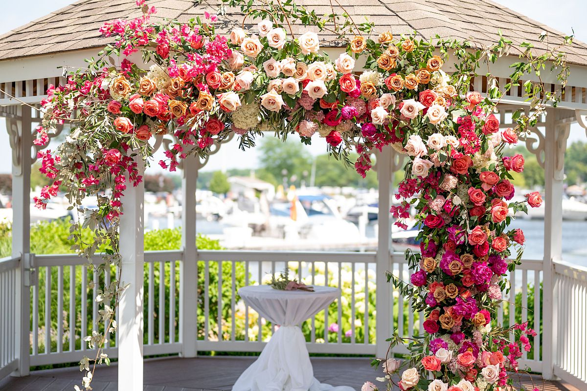 A decorated gazebo with a covering of colorful flowers, possibly set up for a wedding or an outdoor event, located near a body of water.