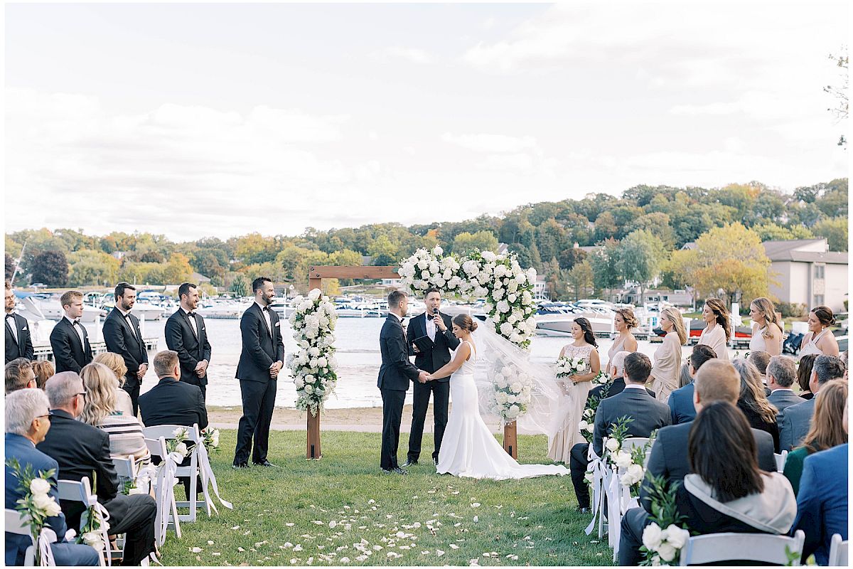 A wedding ceremony by the lake, with a couple under a floral arch, surrounded by guests and wedding party. Everyone is facing the couple.