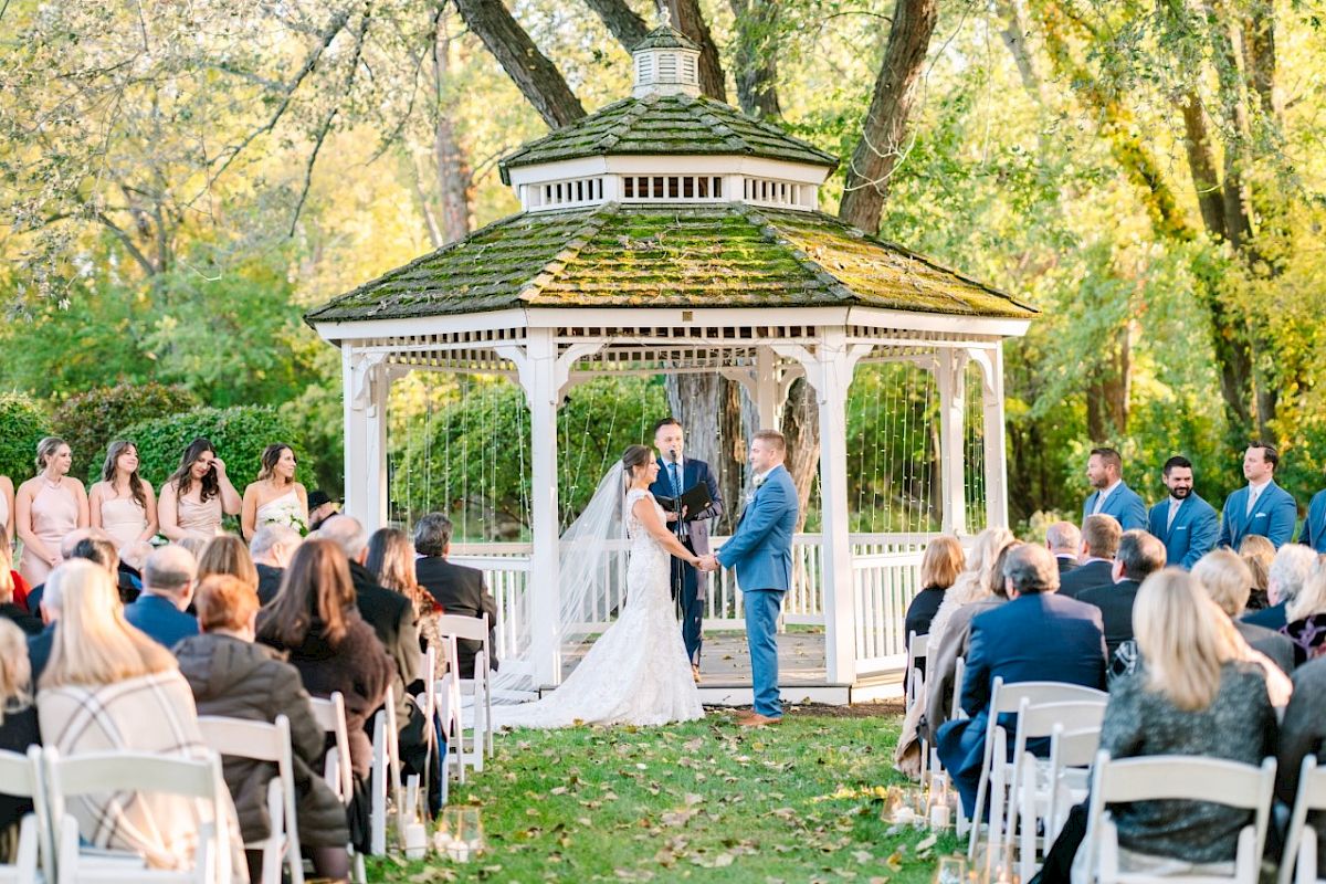 A wedding ceremony is taking place outdoors with a bride and groom standing in front of a white gazebo surrounded by guests.