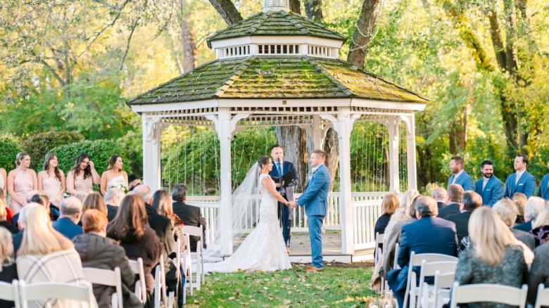 A wedding ceremony is taking place outdoors with a bride and groom standing in front of a white gazebo surrounded by guests.