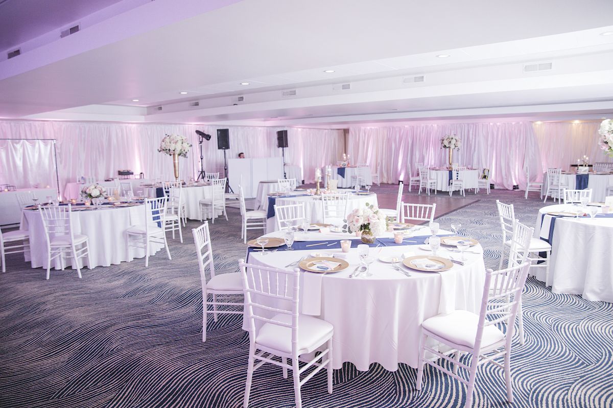 An elegantly set up event space with round tables, white chairs, centerpieces, and ambient lighting. The room is prepared for a wedding or banquet.